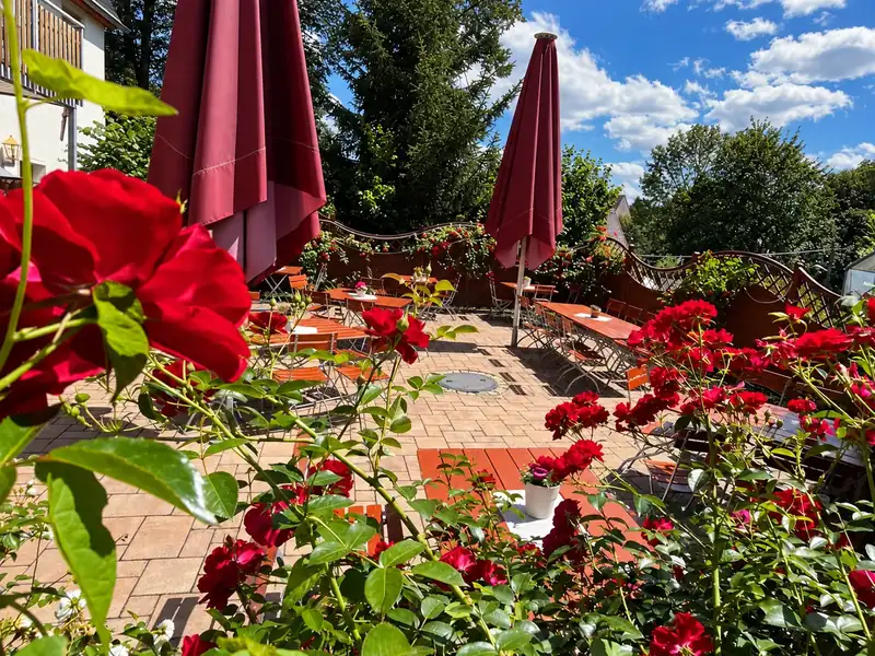 Rote Rosen vor der Terrasse mit Holztischen und Sonnenschirmen